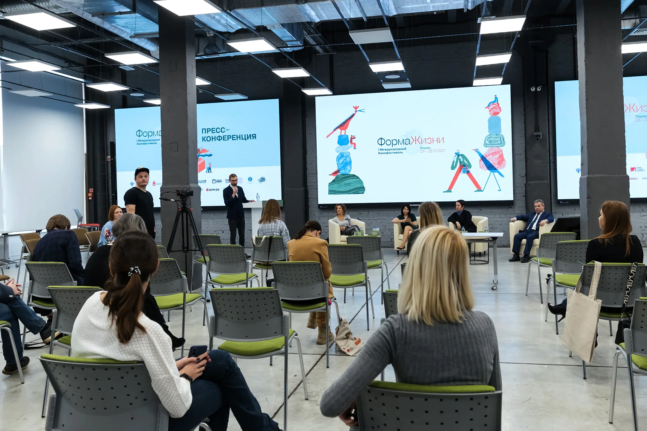 The opening press conference of the first edition: a spaced-out audience on green chairs facing a panel, with the identity running across three LED walls behind them.
