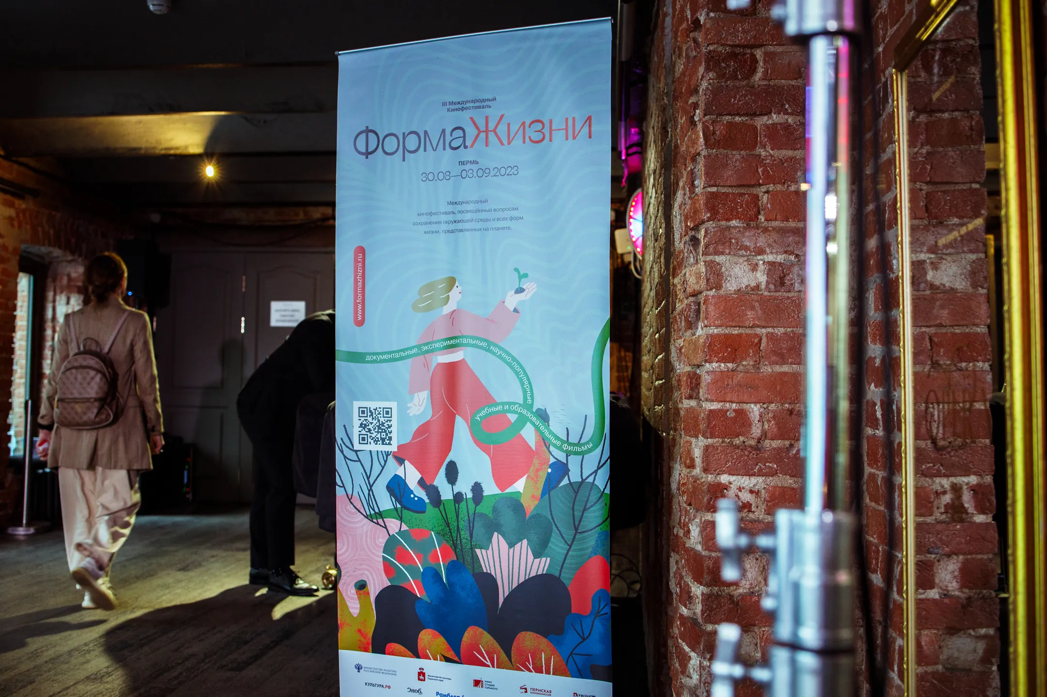 A festival roll-up banner in a dimly lit venue corridor with brick walls, a visitor walking past.