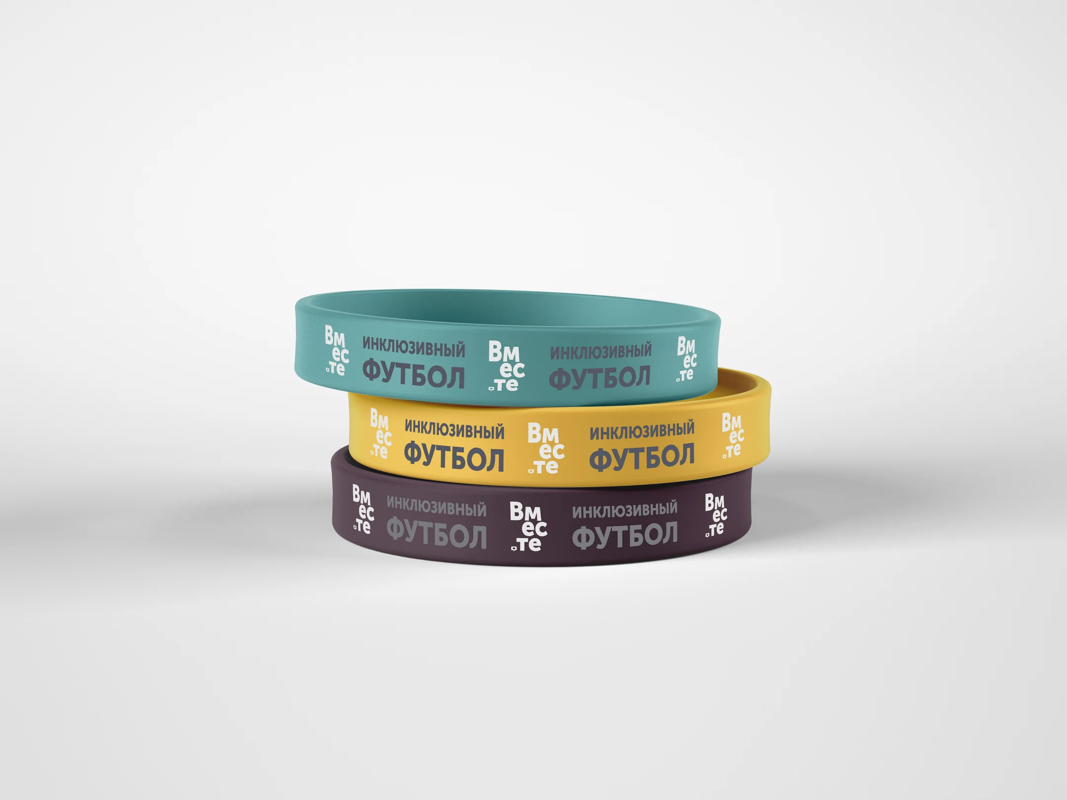 Three stacked silicone wristbands in teal, gold and plum, each embossed with инклюзивный футбол and the Vmeste name.