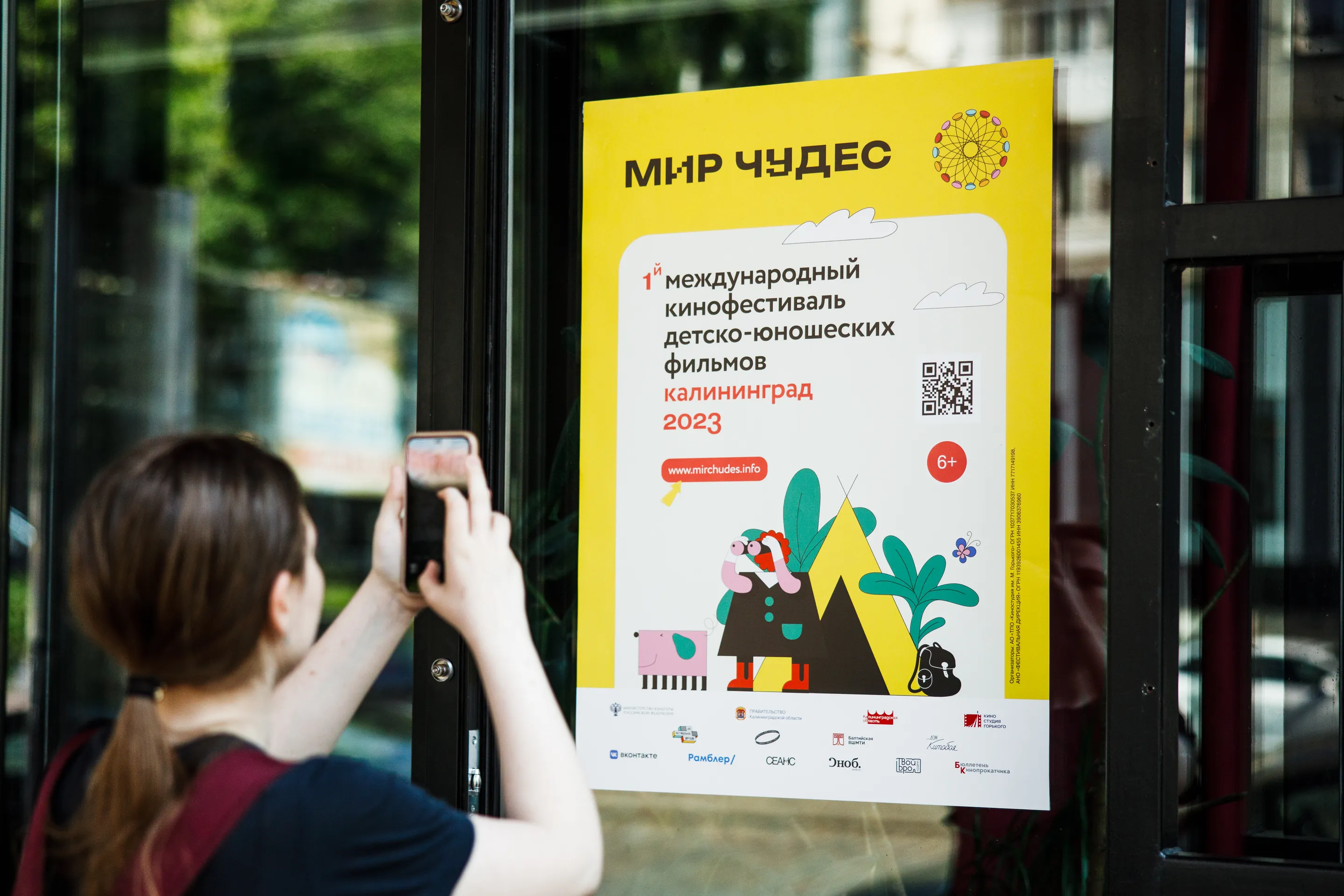 A passer-by photographing the yellow World of Wonders citylight poster on a Kaliningrad street.